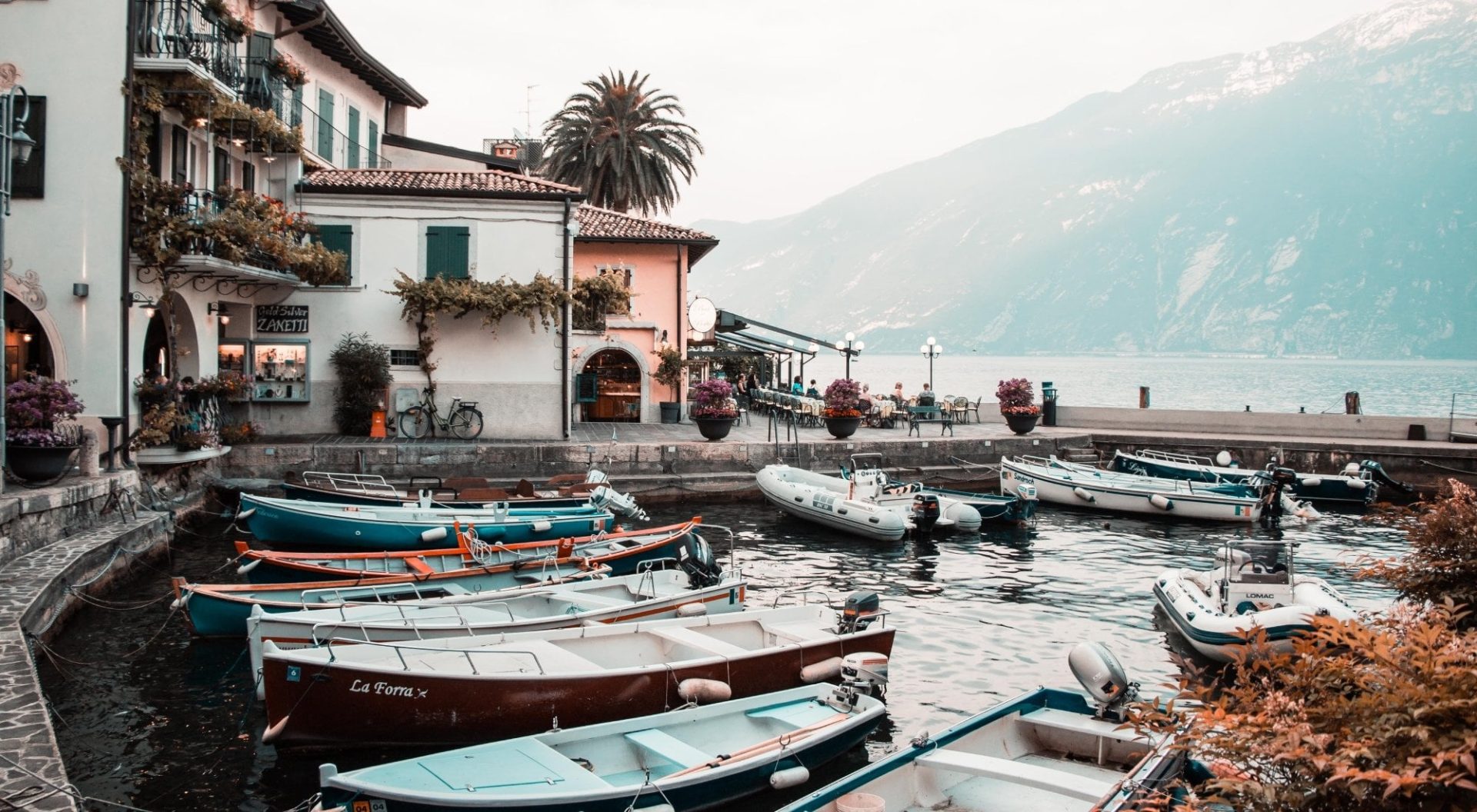 Small harbour on Lake Garda with moored boats, lakeside buildings, and mountain backdrop