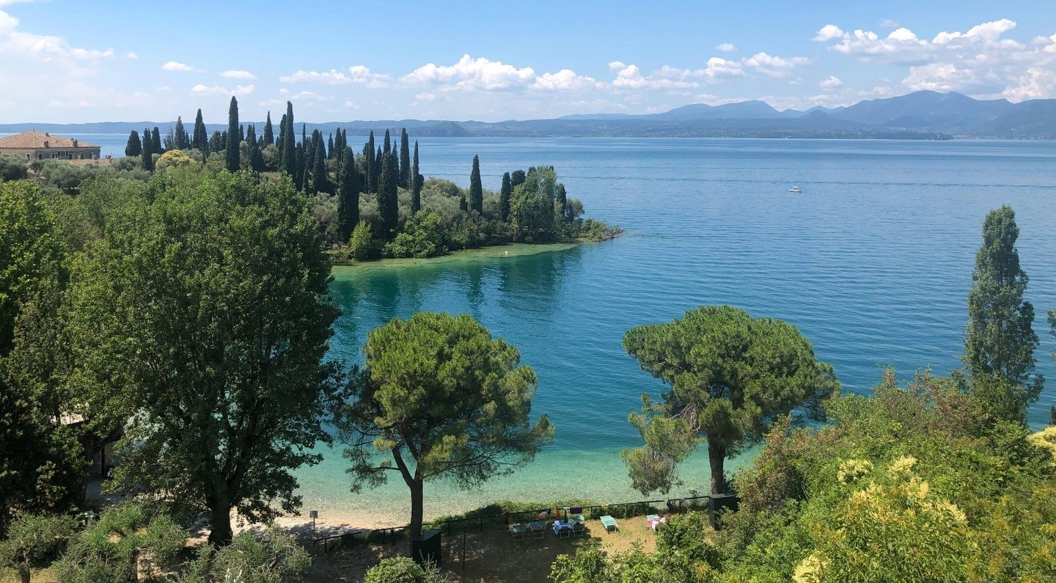 Cypress trees lining the shoreline of Lake Garda with clear blue water and distant mountains
